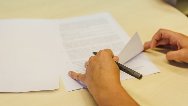 Eine Person hält ein Dokument und schreibt mit einem Stift auf dem Tisch. Im Hintergrund sind ein Ordner und mehrere Blätter sichtbar.