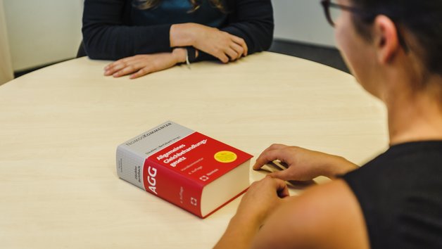 Zwei Personen sitzen an einem Tisch, vor ihnen ein offenes rotes Buch. Beide tragen Casual-Kleidung und unterhalten sich in einem modernen Raum mit neutralem Hintergrund.